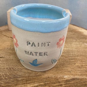 May include: A handmade ceramic paint water cup with a light blue rim and interior. The exterior is off-white with the words "PAINT WATER" and floral and bird designs. The cup is on a wooden surface.