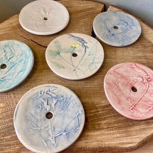 May include: Six ceramic soap dishes with floral patterns. Each round dish has a central drainage hole and a unique colour and botanical design. Colours include white, blue, and pink. The dishes are displayed on a wooden surface.