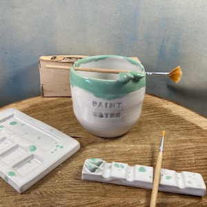 May include: A ceramic artist's set featuring a white and teal paint water cup with the words "PAINT WATER", a white paint palette, and a small brush holder. A wooden box and paintbrushes are also present.