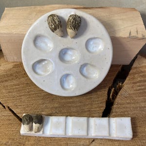 May include: A round, off-white ceramic palette with seven circular indentations and two small, brown-and-white hedgehog figurines attached to the top. Below it is a rectangular, off-white ceramic brush rest with five sections and two matching hedgehog figurines.