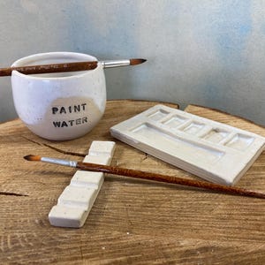 May include: A white ceramic water cup with the text PAINT WATER, a rectangular ceramic paint palette with five wells, a long rectangular ceramic brush rest, and two wooden paint brushes with silver ferrules and brown bristles.