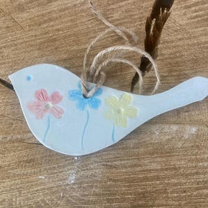 May include: A pale blue ceramic bird decoration featuring three floral designs in pink, blue, and yellow. The bird has a jute string for hanging. The ornament is displayed on a wooden surface.