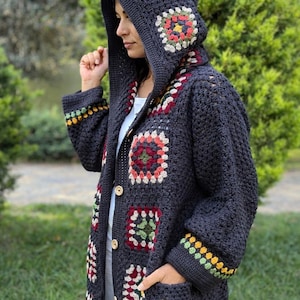May include: A dark gray, hooded crochet cardigan with colorful square patterns. The long-sleeved sweater features a button-up front, pockets, and decorative trim on the cuffs. The hood is also adorned with the same square patterns.