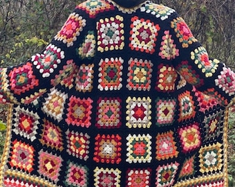 Könnte beinhalten: Eine schwarze Häkeljacke mit einem bunten Granny-Square-Muster. Die Jacke hat einen langen, offenen Vorderteil und einen weiten, fließenden Saum.