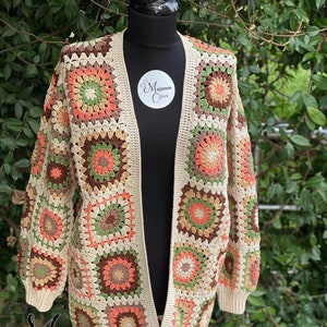 Könnte beinhalten: Ein gehäkelter Cardigan mit einem langen, offenen Vorderteil und langen Ärmeln. Der Cardigan ist aus beigefarbenem Garn gefertigt und hat ein farbenfrohes geometrisches Muster aus Quadraten mit einem Blumendesign. Der Cardigan wird auf einer Puppe präsentiert.