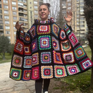 May include: A colorful, handmade crochet poncho with a patchwork design. The poncho features a variety of square patterns in vibrant colors like red, blue, green, and yellow, set against a black background. The person is wearing white sneakers and black leggings.