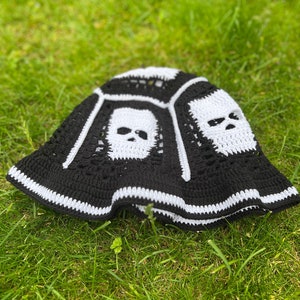 May include: Black and white crocheted bucket hat with skull designs. The hat is lying on green grass.