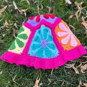 May include: A crocheted hat with a pink brim and a colourful floral design. The hat is made up of six panels, each with a different flower design. The flowers are in shades of pink, orange, yellow, green, blue, and purple.