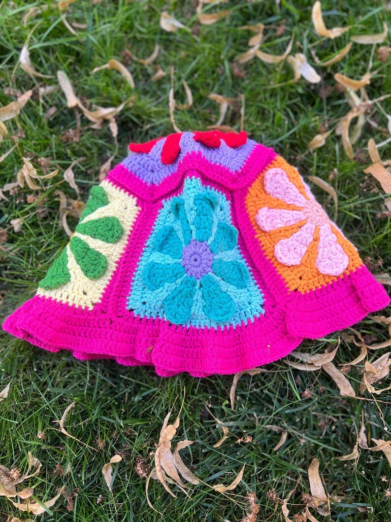 May include: A crocheted hat with a pink brim and a colourful floral design. The hat is made up of six panels, each with a different flower design. The flowers are in shades of pink, orange, yellow, green, blue, and purple.