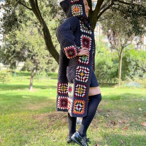 May include: A long, dark blue crocheted cardigan with a hood. The cardigan is decorated with colorful granny squares in shades of red, yellow, green, and white. The cardigan is worn by a person standing in a grassy area.