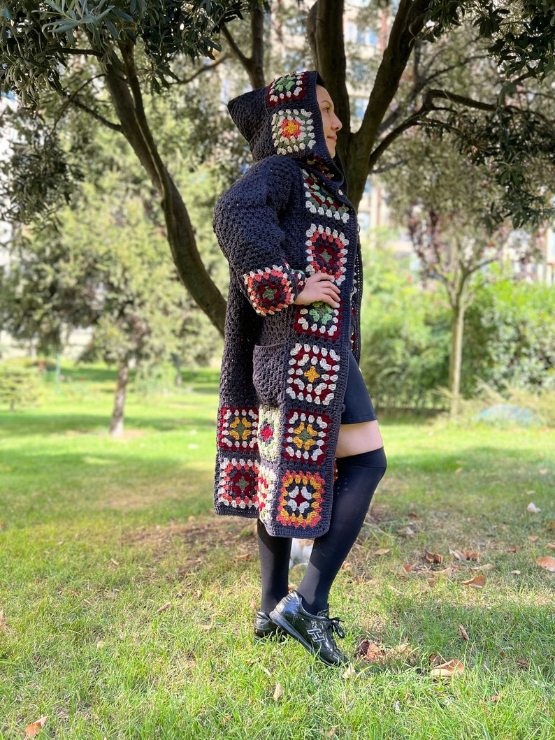 May include: A long, dark blue crocheted cardigan with a hood. The cardigan is decorated with colorful granny squares in shades of red, yellow, green, and white. The cardigan is worn by a person standing in a grassy area.