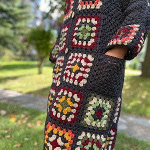 May include: A long, dark gray crochet cardigan with colorful granny square motifs. The cardigan has a button closure and pockets.