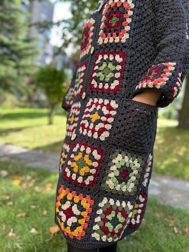 May include: A long, dark gray crochet cardigan with colorful granny square motifs. The cardigan has a button closure and pockets.