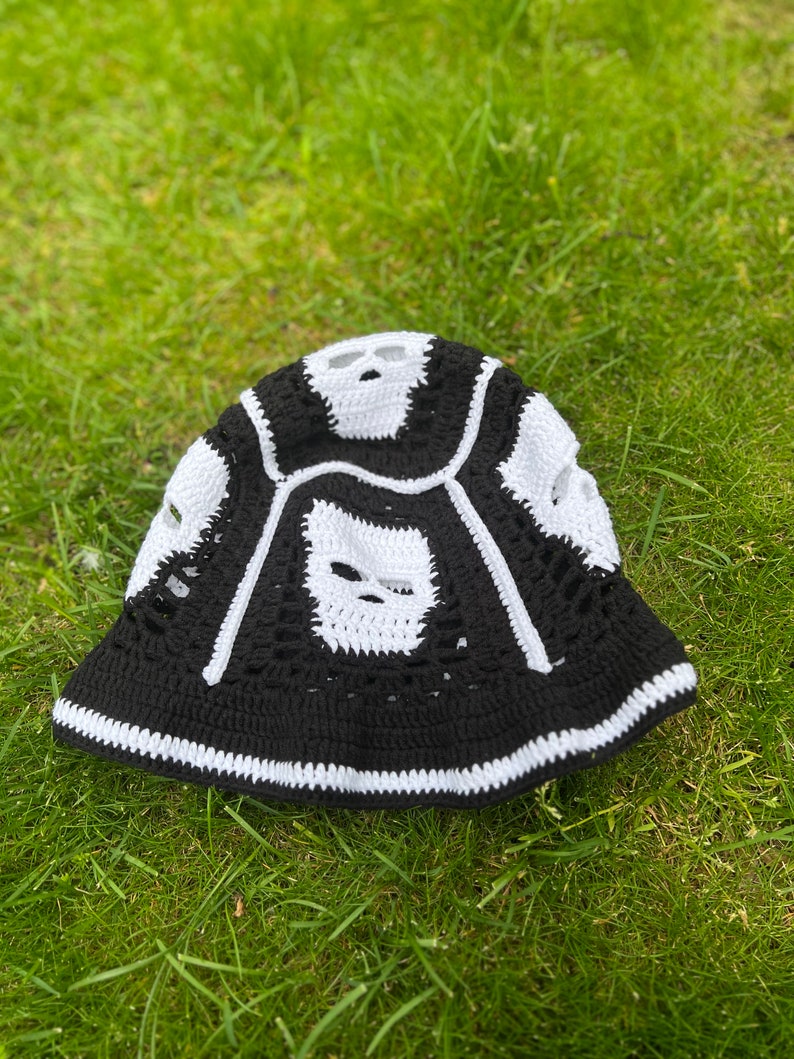 May include: Black and white crocheted bucket hat with a skull design. The hat has a white brim and a black crown with white skull shapes.