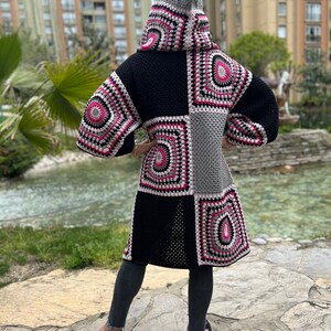 May include: A long, black, gray, and pink crocheted cardigan with a hood. The cardigan has a patchwork design with different sized squares. The cardigan is worn by a person standing on a stone path.