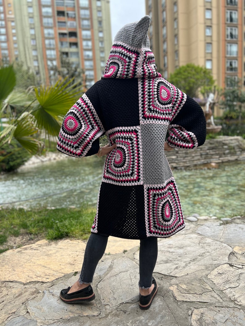 May include: A long, black, gray, and pink crocheted cardigan with a hood. The cardigan has a patchwork design with different sized squares. The cardigan is worn by a person standing on a stone path.