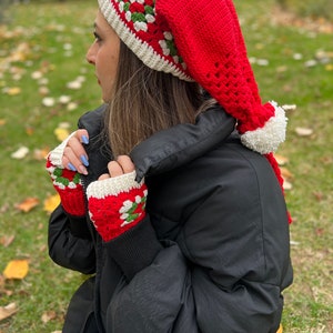 May include: Red and white crocheted hat with a long, pointed, white pom-pom. The hat has a green and white geometric pattern. The hat is worn by a person wearing a black jacket and blue jeans.