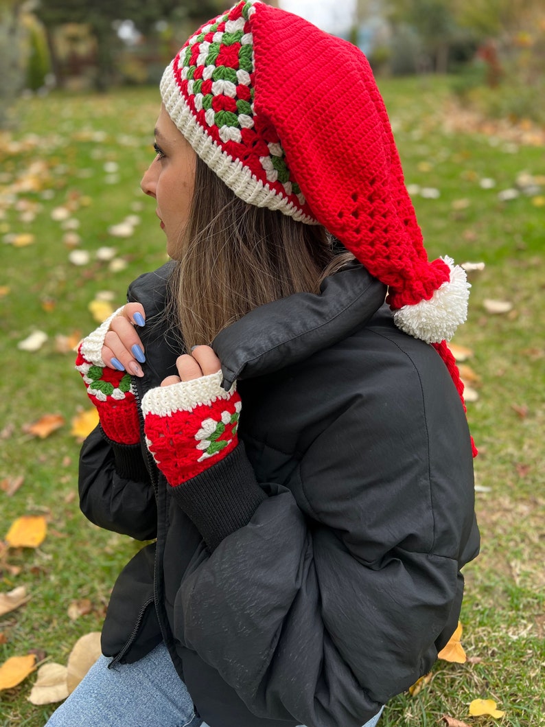 May include: Red and white crocheted hat with a long, pointed, white pom-pom. The hat has a green and white geometric pattern. The hat is worn by a person wearing a black jacket and blue jeans.