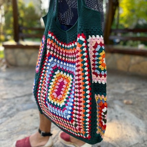 May include: A green crochet tote bag with a colorful granny square design. The bag has a wide, open top and two long straps.