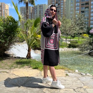 May include: A woman wearing a black and white crocheted cardigan with a pink and white geometric pattern. The cardigan has a hood and is worn over a black top. The woman is wearing white sneakers and sunglasses.