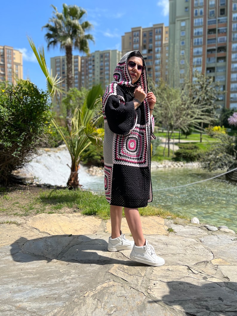 May include: A woman wearing a black and white crocheted cardigan with a pink and white geometric pattern. The cardigan has a hood and is worn over a black top. The woman is wearing white sneakers and sunglasses.