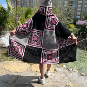May include: A black, gray, and pink crocheted cardigan with a hood. The cardigan is made of squares with different patterns. The cardigan is being worn by a person.
