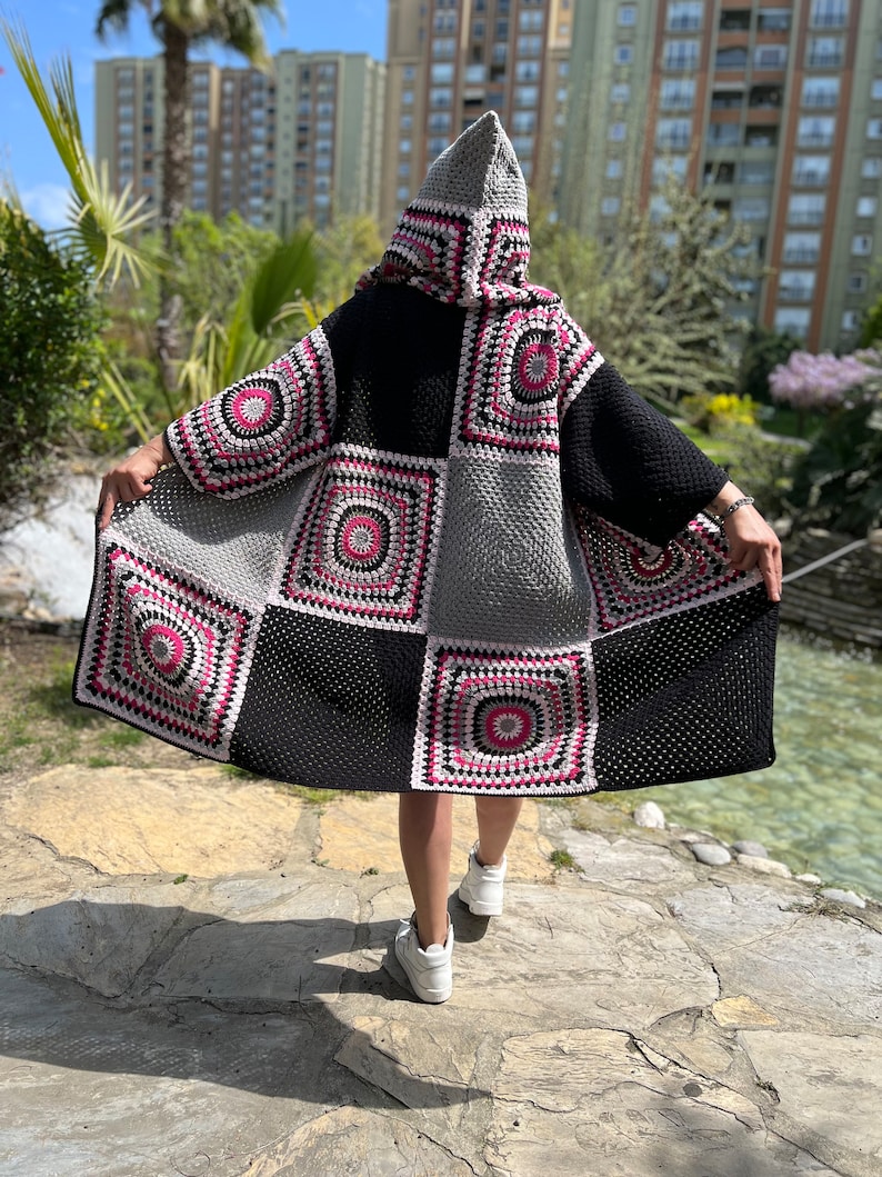 May include: A black, gray, and pink crocheted cardigan with a hood. The cardigan is made of squares with different patterns. The cardigan is being worn by a person.