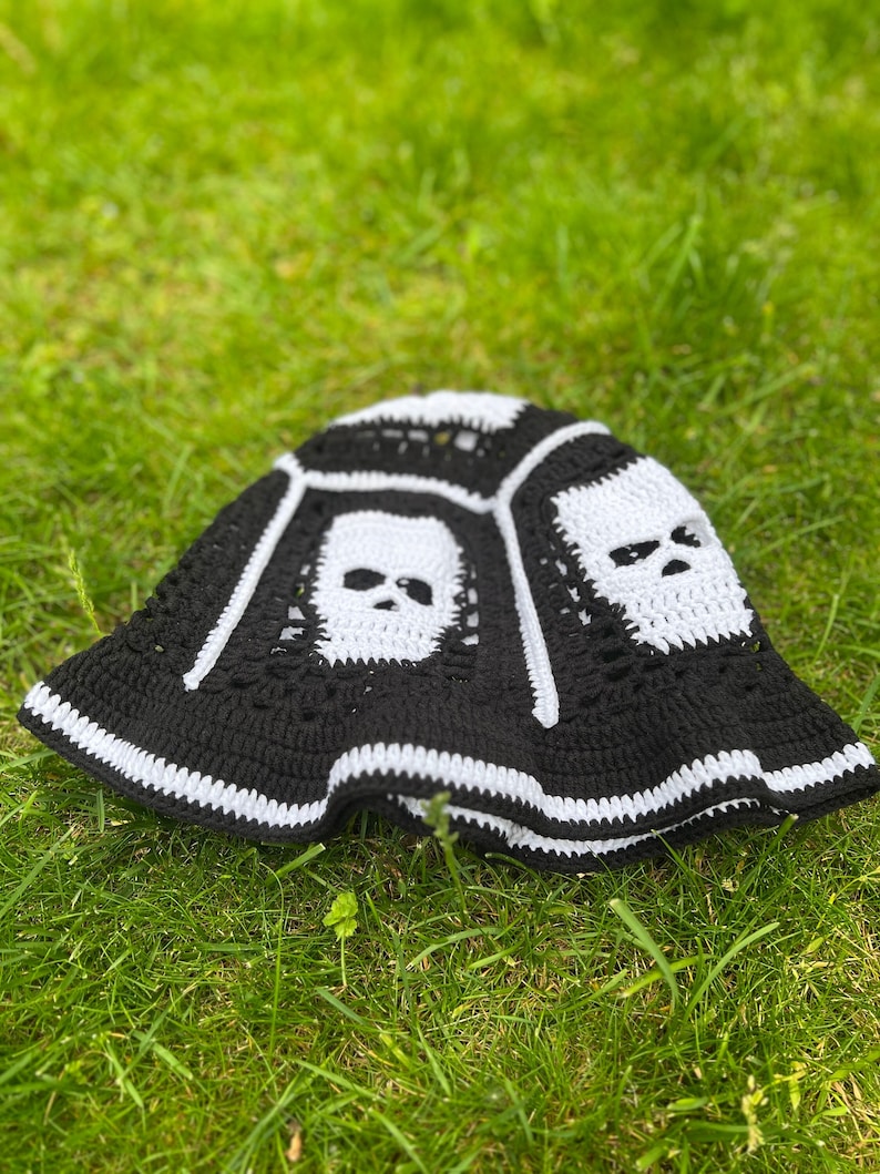 May include: Black and white crocheted bucket hat with skull designs. The hat is lying on green grass.