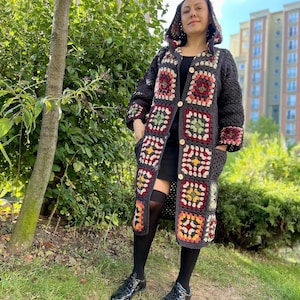 May include: A long, dark gray crocheted cardigan with a hood. The cardigan features a colorful granny square pattern in shades of red, green, yellow, and white. The cardigan has a button closure and pockets.