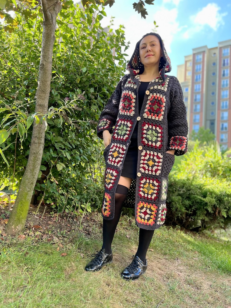 May include: A long, dark gray crocheted cardigan with a hood. The cardigan features a colorful granny square pattern in shades of red, green, yellow, and white. The cardigan has a button closure and pockets.