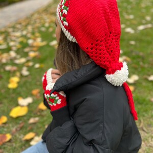 May include: A red crocheted hat with a white pom-pom and green and white trim. The hat has a long tail that hangs down the back. The hat is being worn by a person wearing a black jacket and blue jeans.