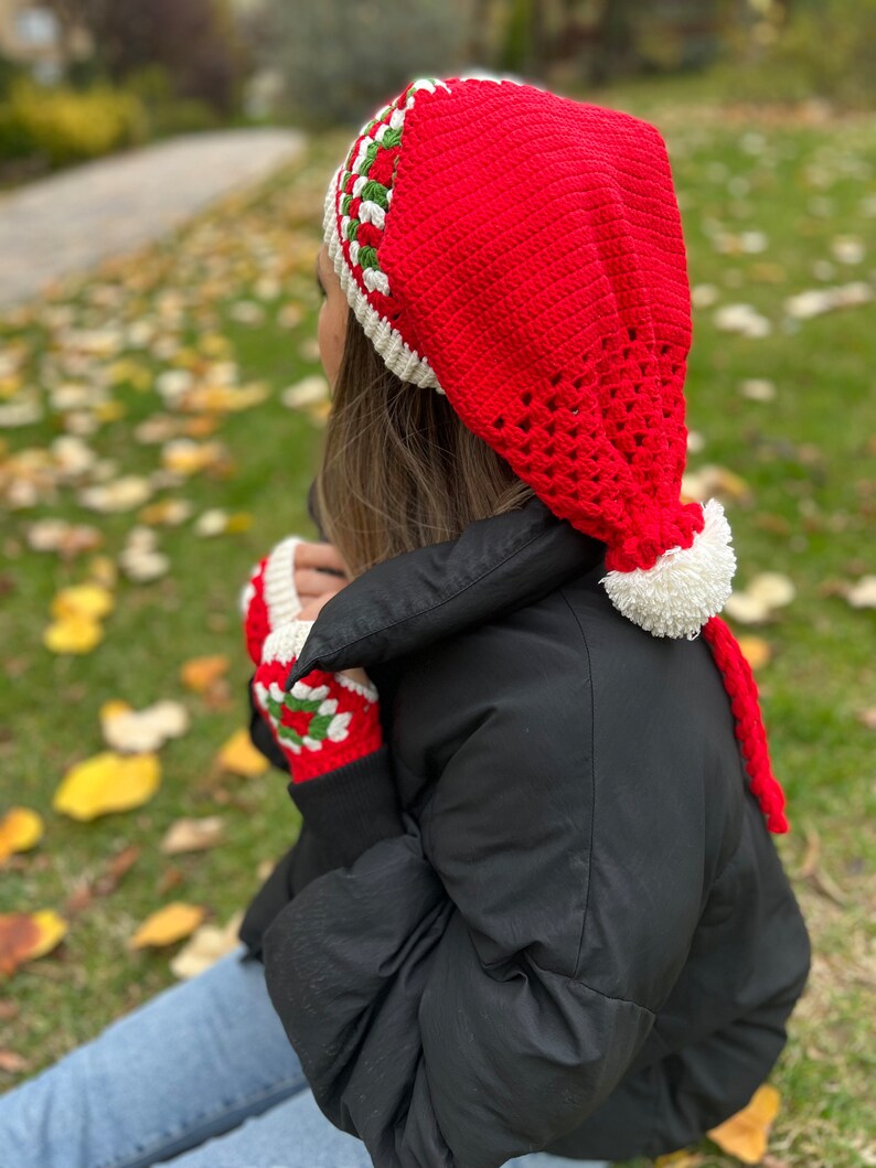 May include: A red crocheted hat with a white pom-pom and green and white trim. The hat has a long tail that hangs down the back. The hat is being worn by a person wearing a black jacket and blue jeans.