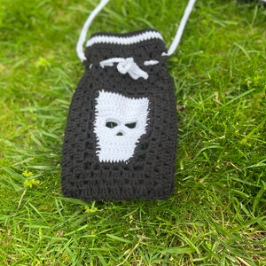 May include: Black and white crocheted bag with a skull design. The bag has a white drawstring and a white strap.