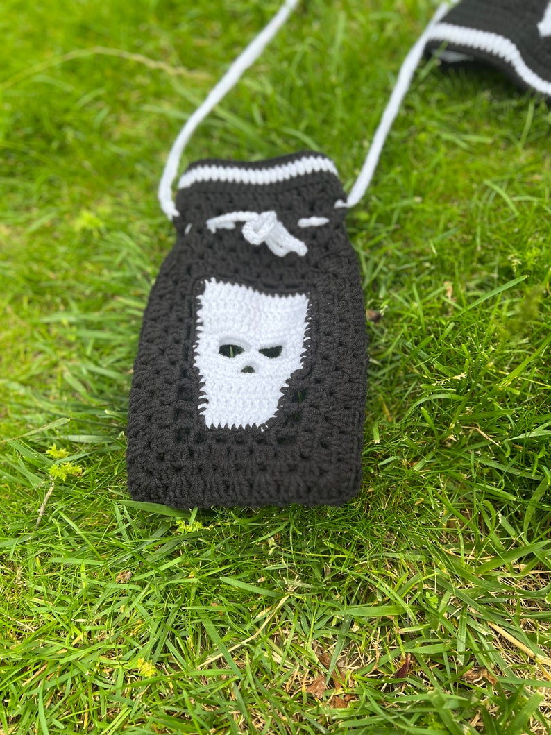 May include: Black and white crocheted bag with a skull design. The bag has a white drawstring and a white strap.