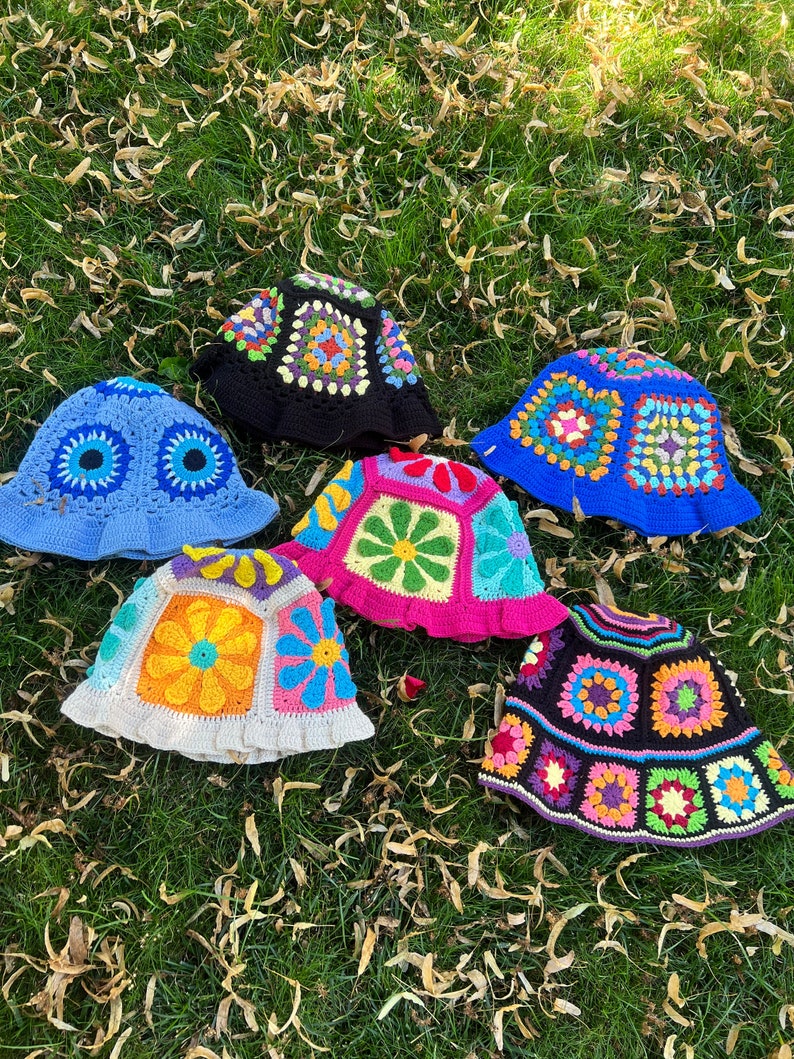 May include: Six crocheted bucket hats in a variety of colours and patterns, including blue, pink, white, and black. The hats are laid out on a bed of green grass with brown leaves.