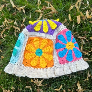 May include: A crocheted bucket hat with a white brim and a patchwork design featuring colourful flowers in shades of yellow, orange, blue, pink, green, and purple.