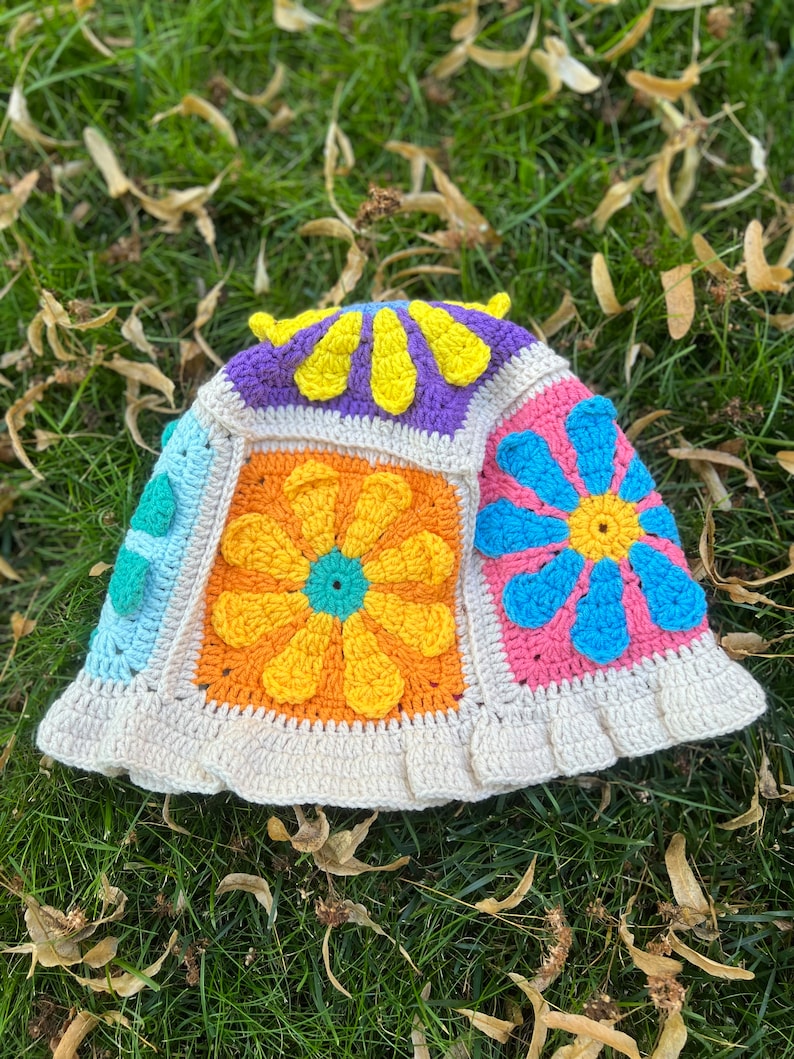 May include: A crocheted bucket hat with a white brim and a patchwork design featuring colourful flowers in shades of yellow, orange, blue, pink, green, and purple.
