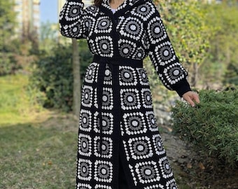May include: A long, black and white crochet cardigan with a geometric pattern. The cardigan has a hood, long sleeves, and a belt. The person is wearing black pants and white sneakers. The background is a park with trees and greenery.