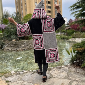 May include: A long black and gray crocheted cardigan with a hood. The cardigan has a patchwork design with pink and white accents.