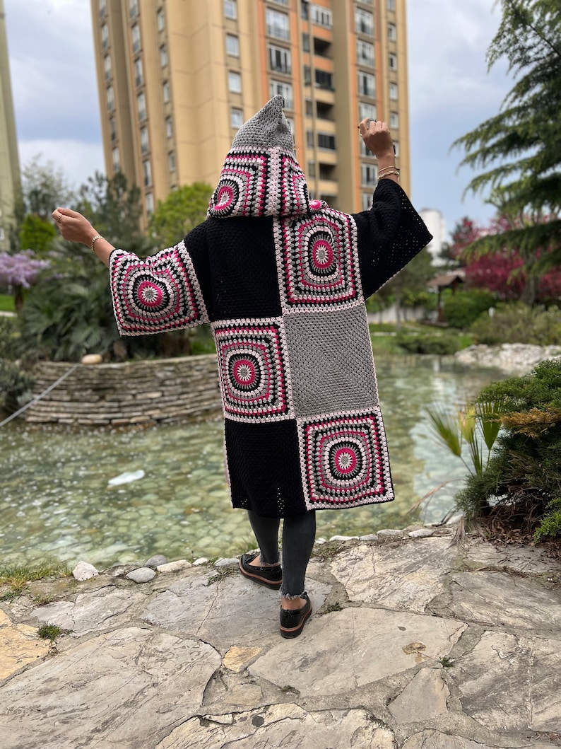 May include: A long black and gray crocheted cardigan with a hood. The cardigan has a patchwork design with pink and white accents.