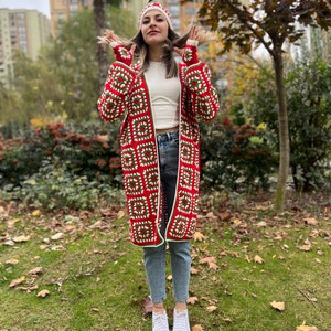 May include: A red and green crochet cardigan with white accents. The cardigan is long and has a hood. The model is wearing the cardigan with a white crochet hat and blue jeans.
