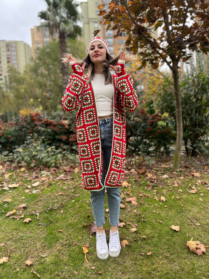 May include: A red and green crochet cardigan with white accents. The cardigan is long and has a hood. The model is wearing the cardigan with a white crochet hat and blue jeans.