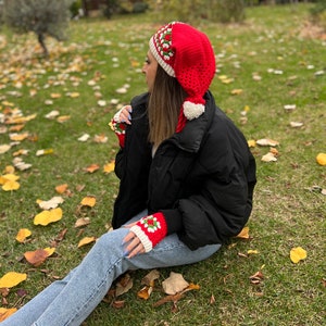 May include: A red and white crocheted hat with a pom-pom on top. The hat is shaped like a Santa hat with a long pointed tip. The hat is worn by a person sitting on a grassy surface with fallen leaves.