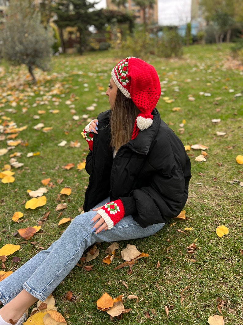 May include: A red and white crocheted hat with a pom-pom on top. The hat is shaped like a Santa hat with a long pointed tip. The hat is worn by a person sitting on a grassy surface with fallen leaves.