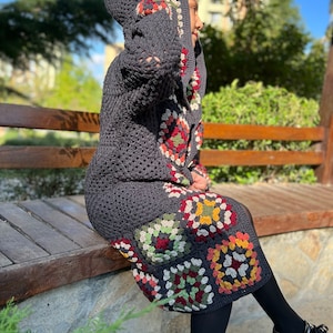 May include: A gray crocheted hooded cardigan with a colorful granny square pattern. The cardigan is worn over a black skirt and black tights.