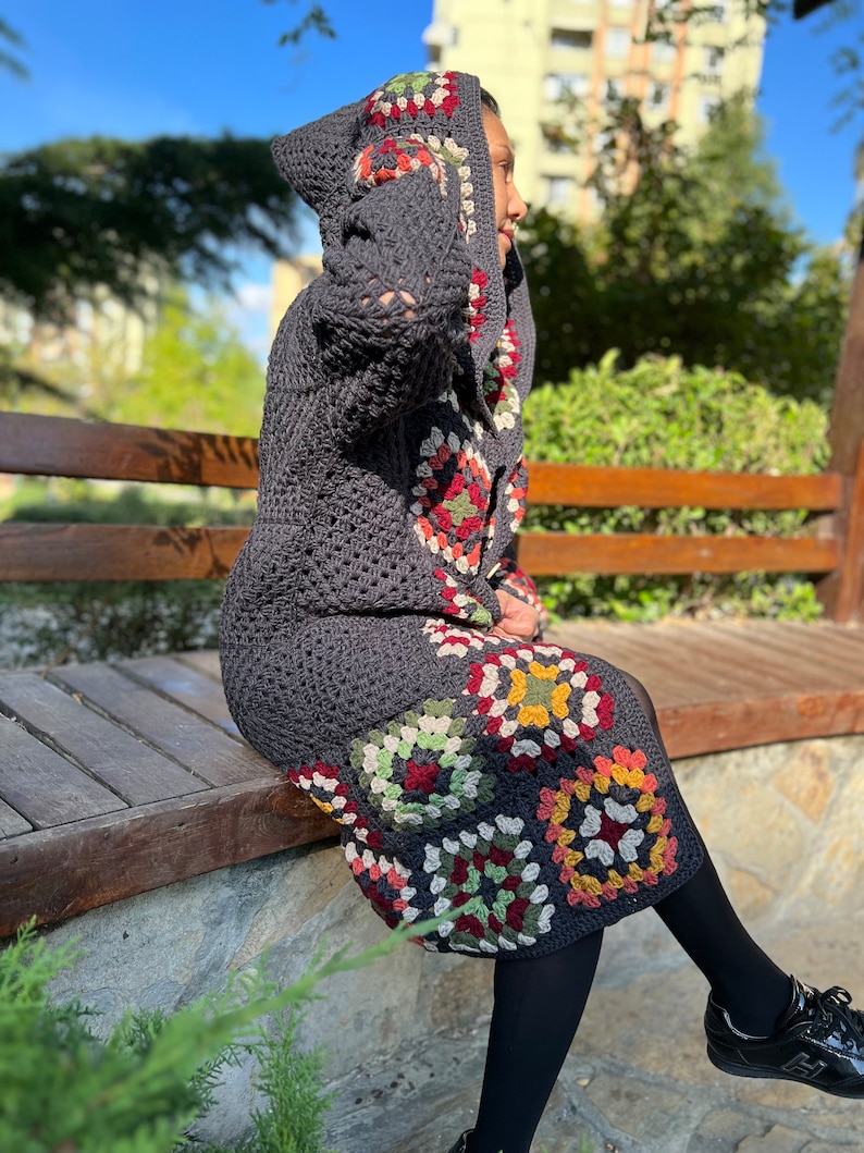 May include: A gray crocheted hooded cardigan with a colorful granny square pattern. The cardigan is worn over a black skirt and black tights.