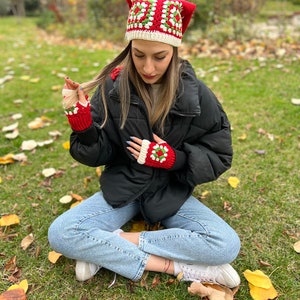 May include: A red and green crocheted hat with a white border. The hat has a square pattern and is worn by a person sitting on the grass. The person is also wearing red and green crocheted fingerless gloves.