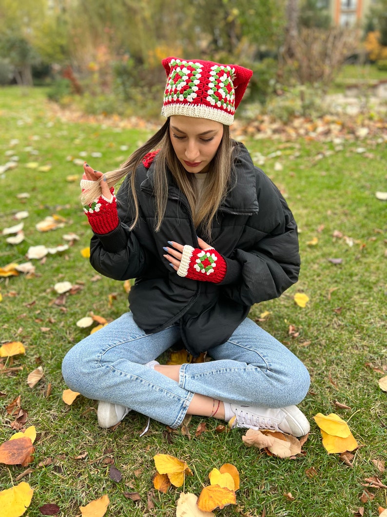 May include: A red and green crocheted hat with a white border. The hat has a square pattern and is worn by a person sitting on the grass. The person is also wearing red and green crocheted fingerless gloves.