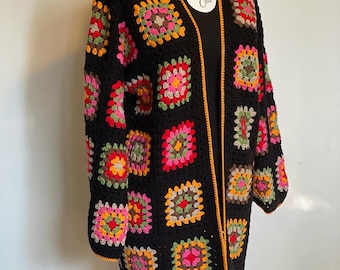 Könnte beinhalten: Eine schwarze Häkeljacke mit einem bunten Granny-Square-Muster. Die Jacke hat einen langen, offenen Vorderteil und einen gelben Saum.