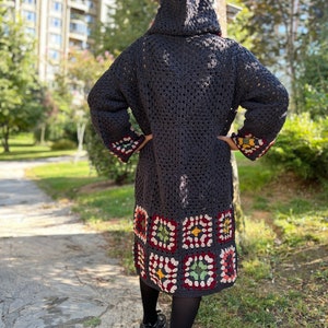May include: A long, dark gray crocheted cardigan with a hood. The cardigan has a colorful geometric pattern at the bottom hem.